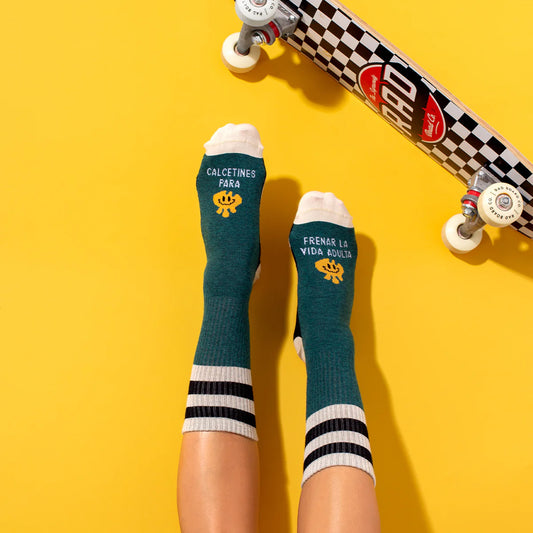 Pies con los calcetines verdes con una carita amarilla sonriente y el mensaje "Calcetines para frenar la vida adulta" con un skate y sobre fondo amarillo