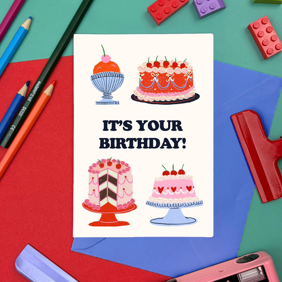 Minitarjeta It's Your BDay de Laura Ortiz, con tartas y el mensaje «It's your birthday!», sobre un sobre azul entre lápices y papelería.
