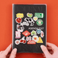 Portada de la Agenda 2027 negra Journal Sessions personalizada con pegatinas incluidas: dados, cerezas, gato, vaca, frases como No todo es urgente y Me lo permito, sostenida por dos manos sobre fondo naranja