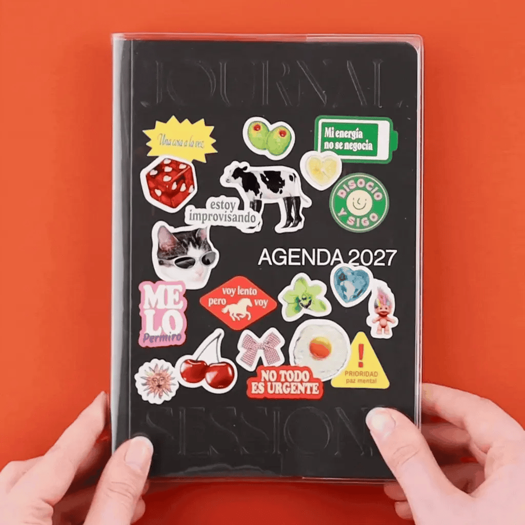 Portada de la Agenda 2027 negra Journal Sessions personalizada con pegatinas incluidas: dados, cerezas, gato, vaca, frases como No todo es urgente y Me lo permito, sostenida por dos manos sobre fondo naranja