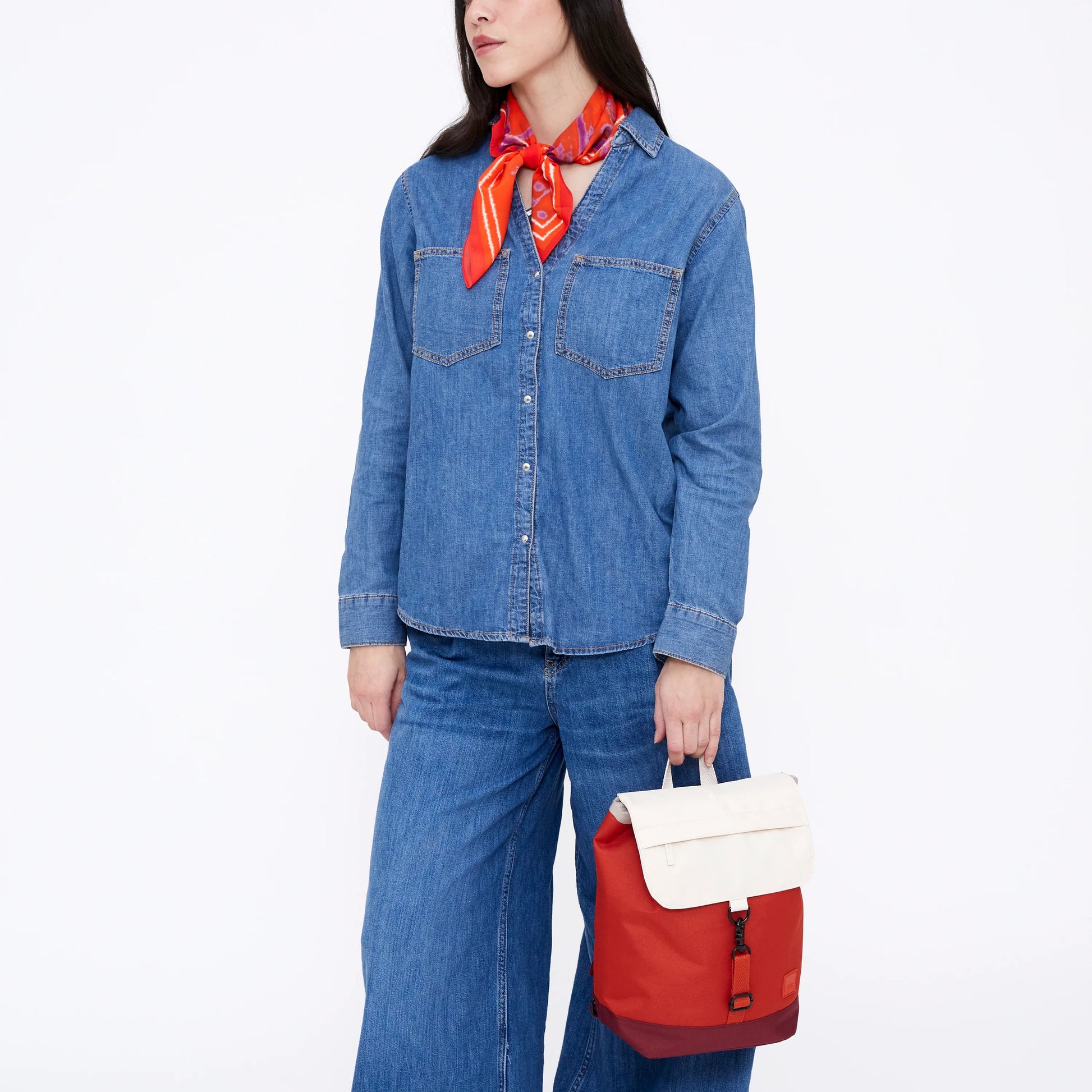 Mujer de frente vestida con pantalón y camisa vaqueros, pañuelo rojo y una mochila color cereza y blanca en la mano