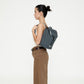 Modelo de perfil llevando la mini mochila Lefrik Kaut color laurel a la espalda, con camiseta blanca y pantalón marrón