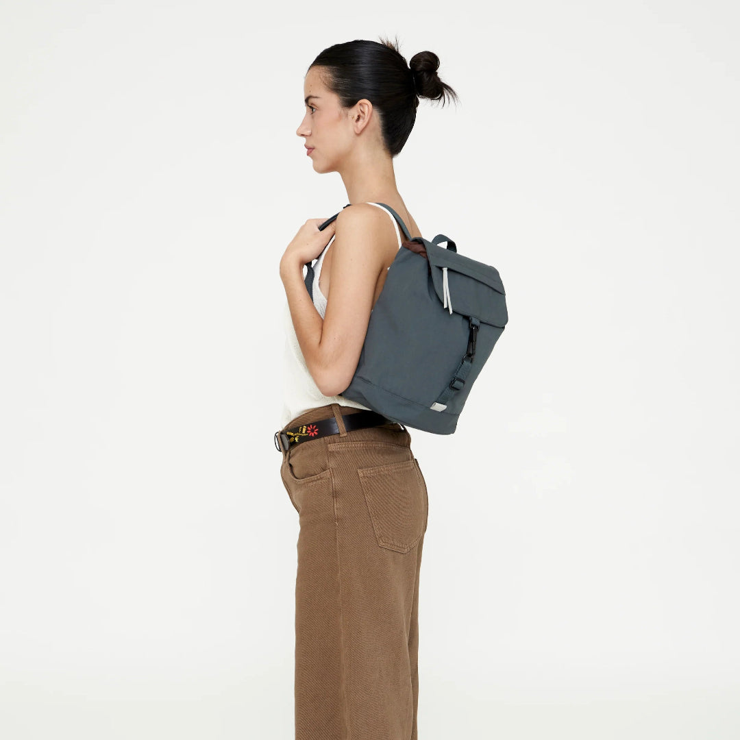Modelo de perfil llevando la mini mochila Lefrik Kaut color laurel a la espalda, con camiseta blanca y pantalón marrón