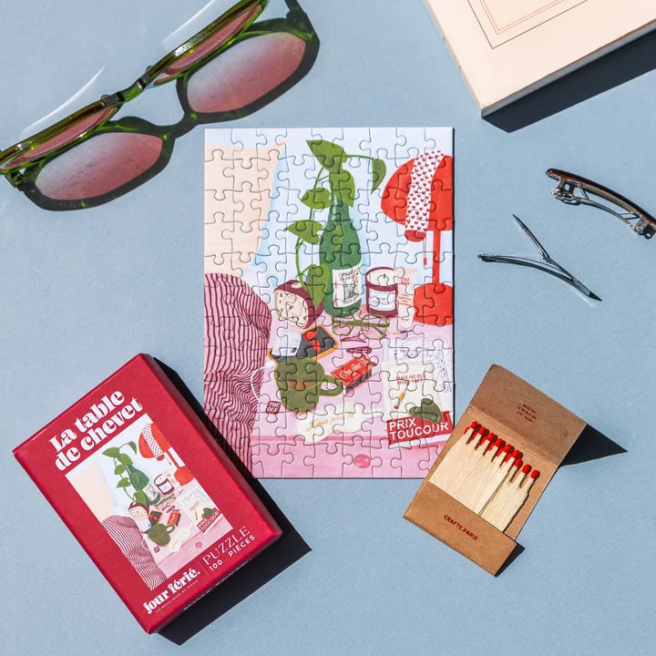 Puzzle La Table de Chevet a medio montar junto a su caja roja, unas gafas de sol y una caja de cerillas sobre fondo azul, de Jour Férié
