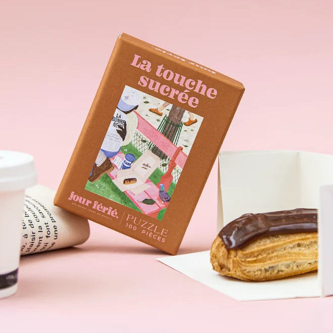 Caja marrón del puzzle La Touche Sucrée de 100 piezas de Jour Férié, apoyada junto a un café para llevar y un éclair de chocolate sobre fondo rosa