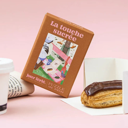 Caja marrón del puzzle La Touche Sucrée de 100 piezas de Jour Férié, apoyada junto a un café para llevar y un éclair de chocolate sobre fondo rosa