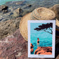 Láminas A5 con ilustración de una chica de espaldas en bañador negro sobre una roca mirando al Mar Mediterráneo apoyada sobre un sombrero de paja a la orilla del mar