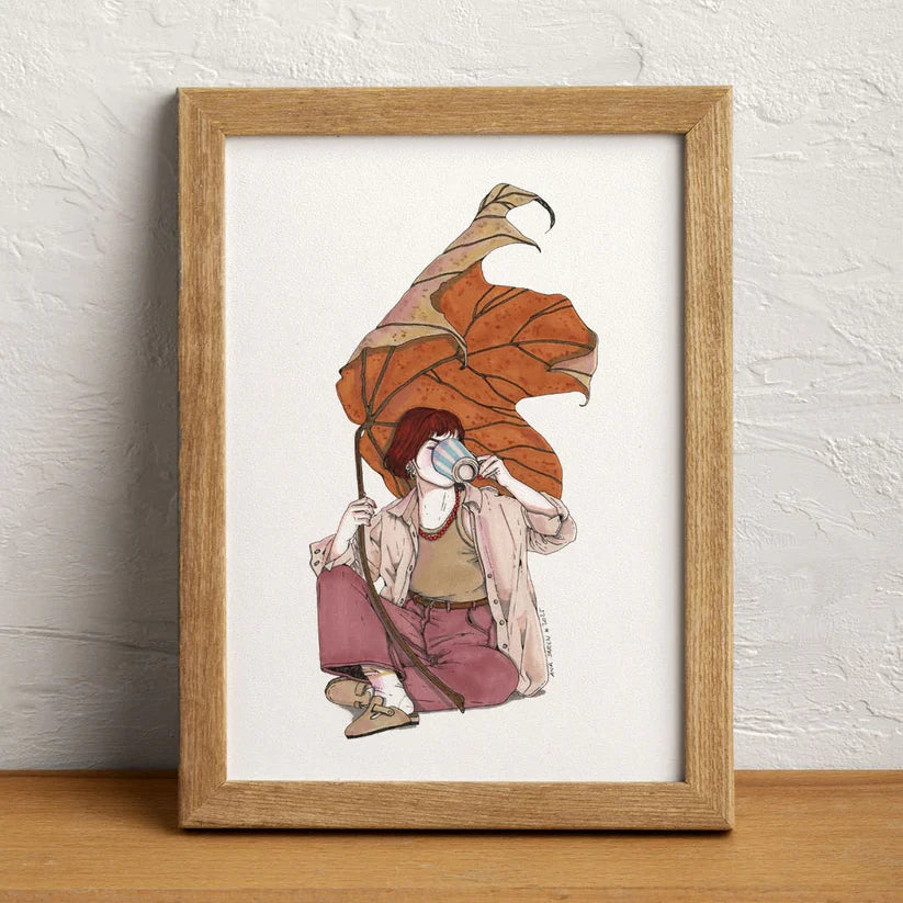 Ilustración enmarcada en un marco de madera de Ana Jarén deuna chica sentada en el suelo bebiendo de un mug a rayas y tapada con una hoja seca