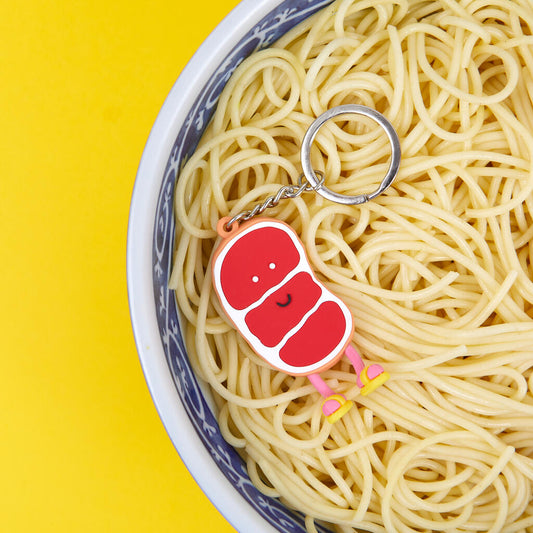 Parte frontal de llavero de filete de carne de ternera Gyu Sensei, roja con detalles blancos que imitan la grasa de la propia carne, tiene una cara sonriente, piernas rosas y chanclas amarillas, reposa sobre un bol de fideos, sobre fondo amarillo. 