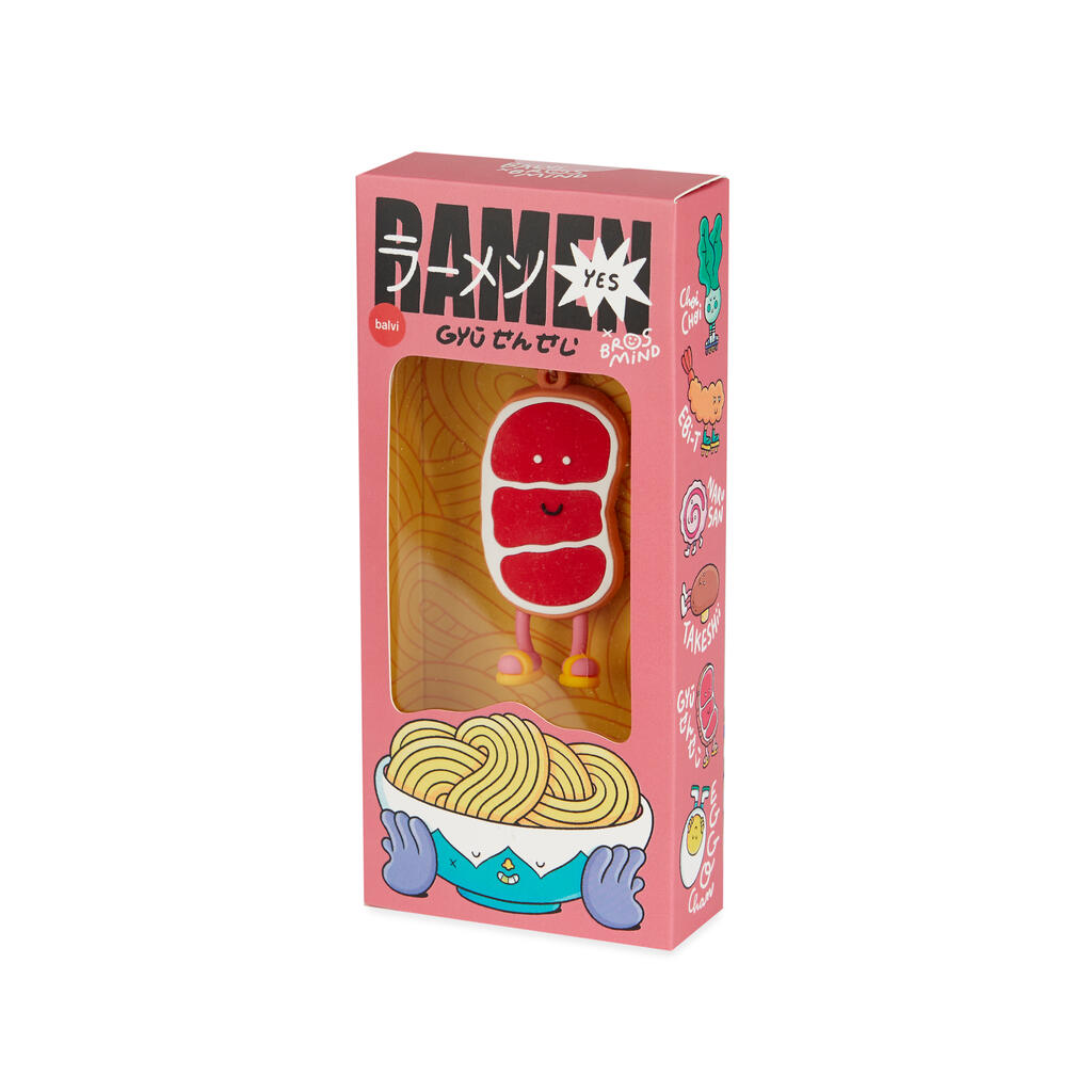 Caja rosa con diferentes dibujos de ingredientes de ramen en ella, dentro vemos la parte frontal de llavero de filete de carne de ternera Gyu Sensei, roja con detalles blancos que imitan la grasa de la propia carne, tiene una cara sonriente, piernas rosas y chanclas amarillas, sobre fondo blanco. 