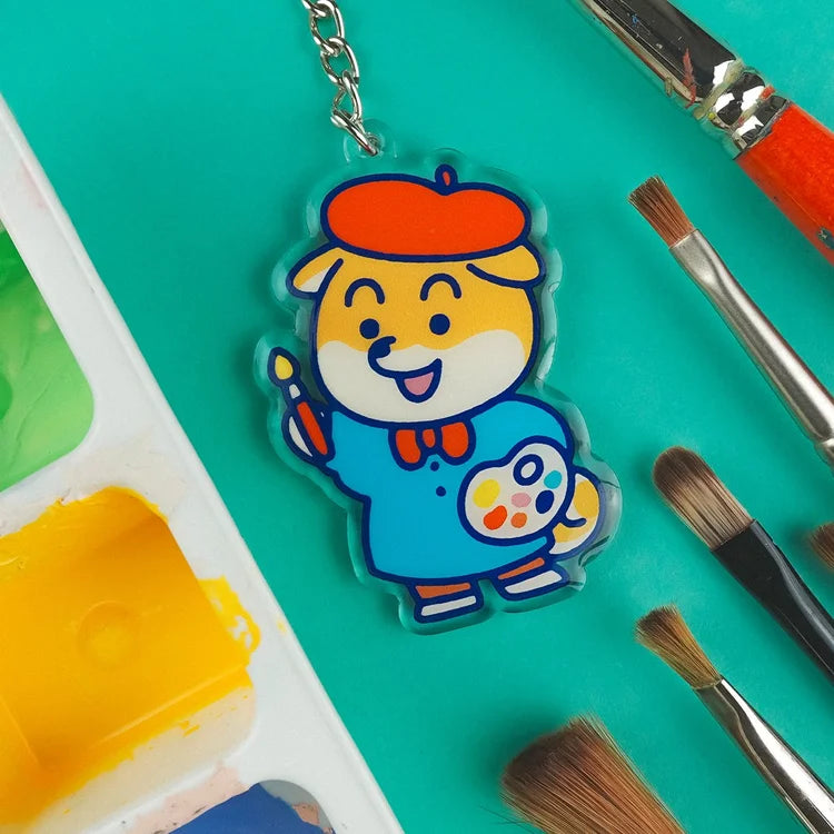 Llavero de acrílico reciclado con forma de perro shiba con boina de artista, pincel y paleta de acuarelas rodeado de pinceles reales