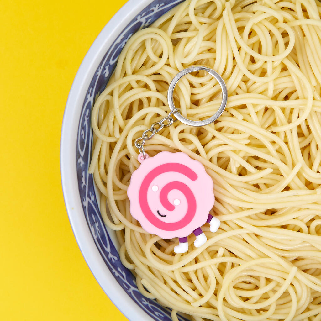 Parte frontal de un naruto, un circulo rosa con una espiral fucsia dentro, y carita sonriente, de tres patas reposando sobre unos fideos dentro de un bol blanco y azul sobre fondo amarillo. 