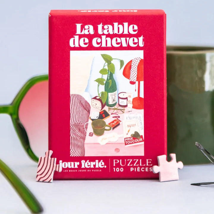 Caja roja del puzzle La Table de Chevet de 100 piezas de Jour Férié, rodeada de gafas de sol verdes, una taza y un clip sobre fondo gris azulado