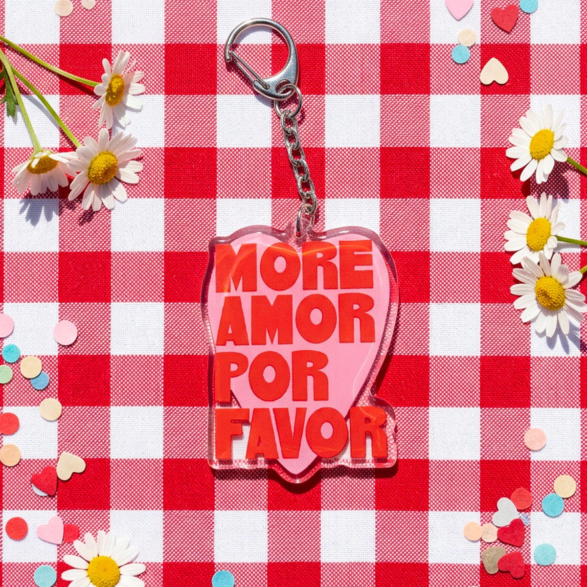 Llavero More Amor de Laura Ortiz, con corazón rosa y letras rojas «More amor por favor», sobre mantel de cuadros con margaritas.
