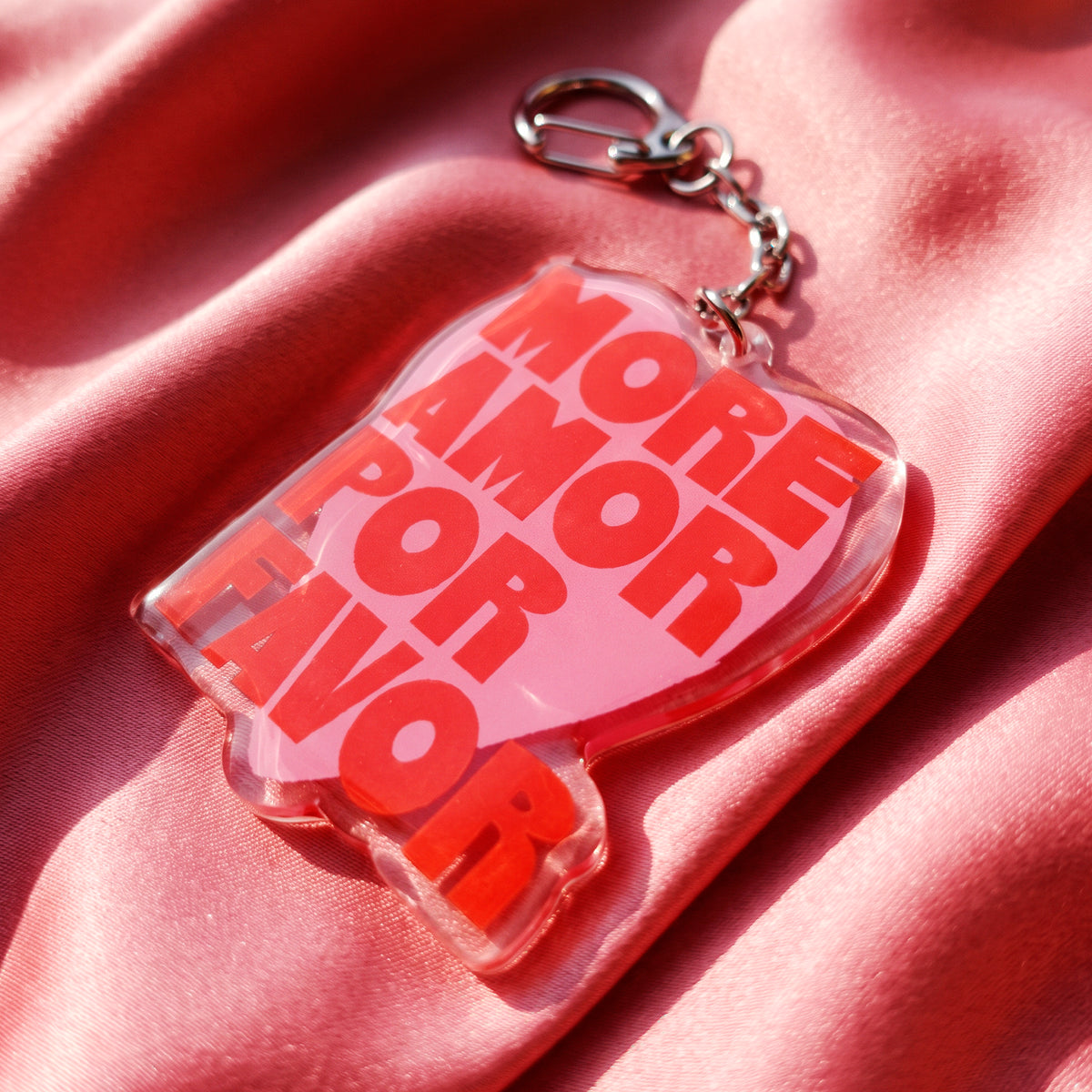 Detalle del llavero acrílico More Amor de Laura Ortiz, con corazón rosa, letras rojas y cierre metálico, sobre tela rosa.
