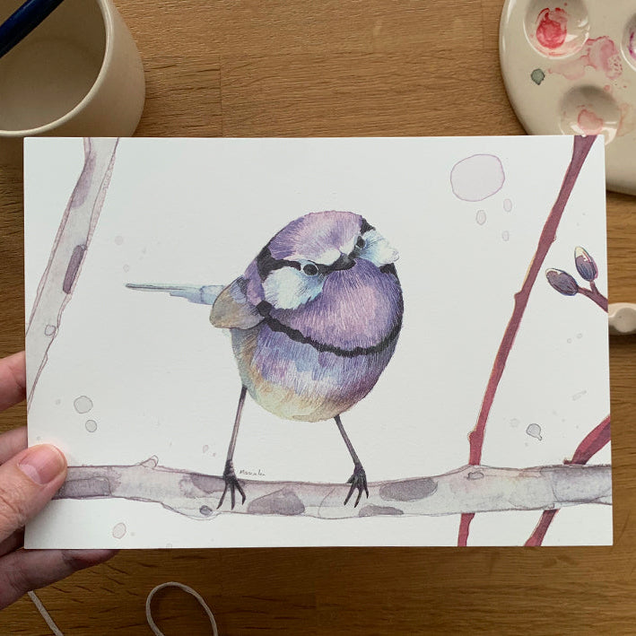 Mini print con ilustración de un pájaro morado de Marialu Gili fotografiada sobre una mesa con acuarelas