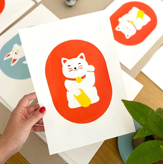 Mano sujetando una print de estilo japonés con un Maneki Neko rojoblanco sobre fondo rojo. Al fondo se ven otras ilustraciones con motivos japoneses. Una planta aporta un toque natural a la escena.