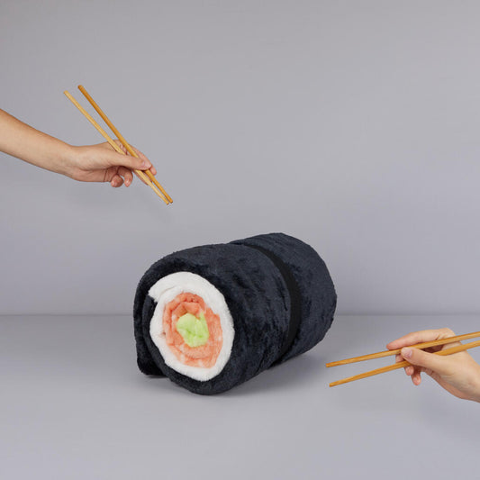Manta Maki negro con forma de roll de sushi sobre mesa gris, con dos manos sujetando palillos japoneses
