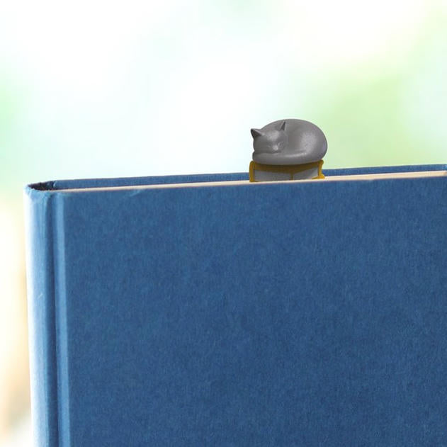 En un libro azul cerrado se asoma un marcapáginas 3D de un libro amarillo con un gato gris dormido encima