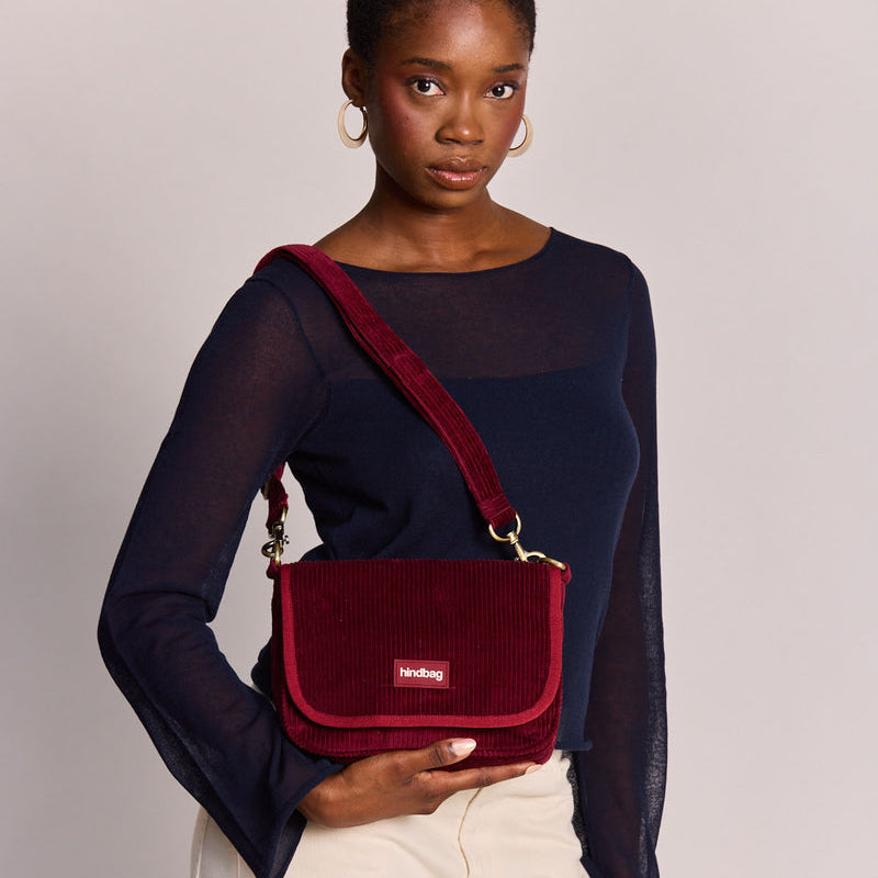 Mujer con camiseta azul y pantalones blancos con un bolso cuadrado con solapa de pana rojo burdeos de la marca francesa Hindbag