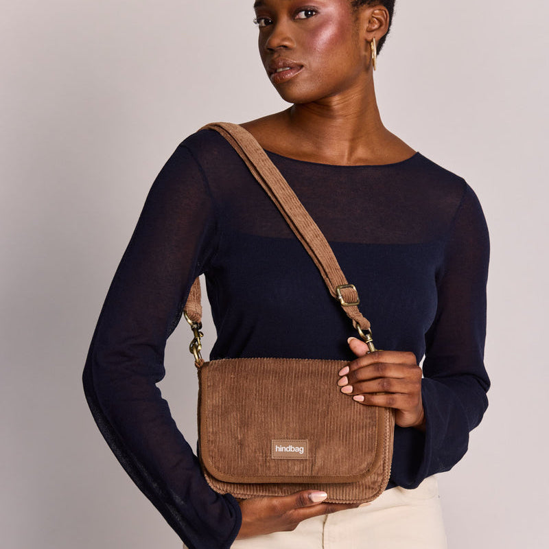 Mujer con camiseta azul y pantalones blancos con un bolso cuadrado con solapa de pana marrón café con leche de la marca francesa Hindbag