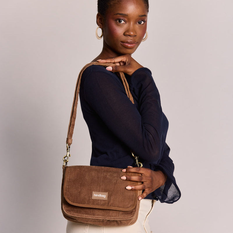 Mujer de perfil con camiseta azul y pantalones blancos con un bolso cuadrado con solapa de pana marrón café con leche de la marca francesa Hindbag colgado al hombro