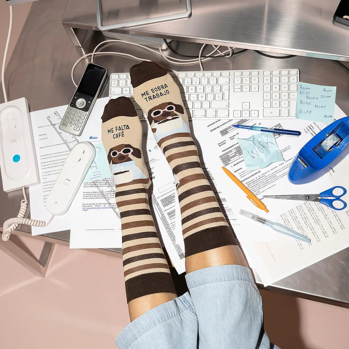 Calcetines de UO con rayas beige y marrones y el mensaje «Me falta café, me sobra trabajo», puestos con los pies sobre un escritorio.