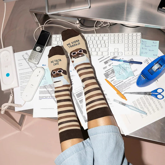 Calcetines de UO con rayas beige y marrones y el mensaje «Me falta café, me sobra trabajo», puestos con los pies sobre un escritorio.