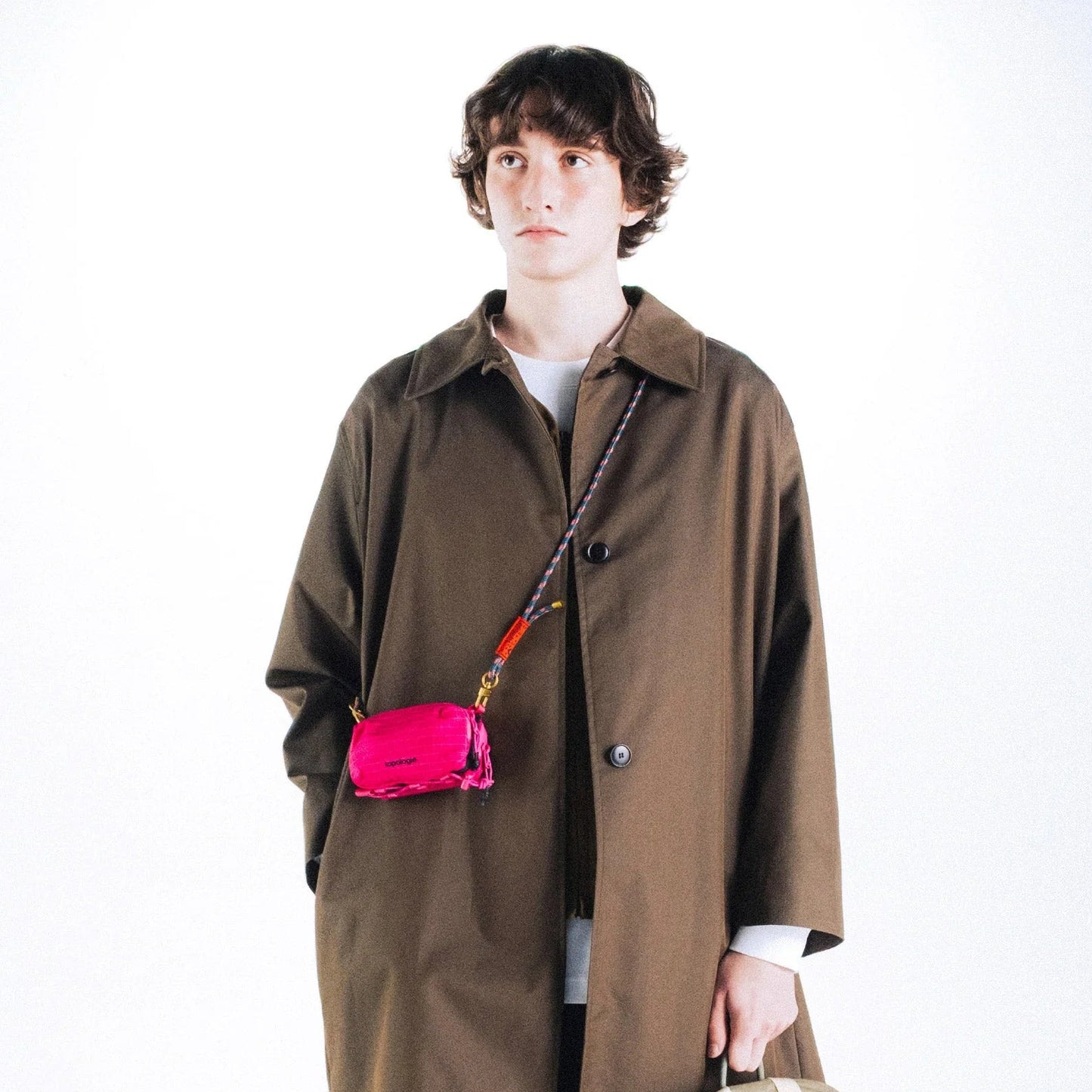 Chico con abrigo marrón y un  bolso Mini Bottle Sacoche de Topologie en color fucsia colgado en bandolera