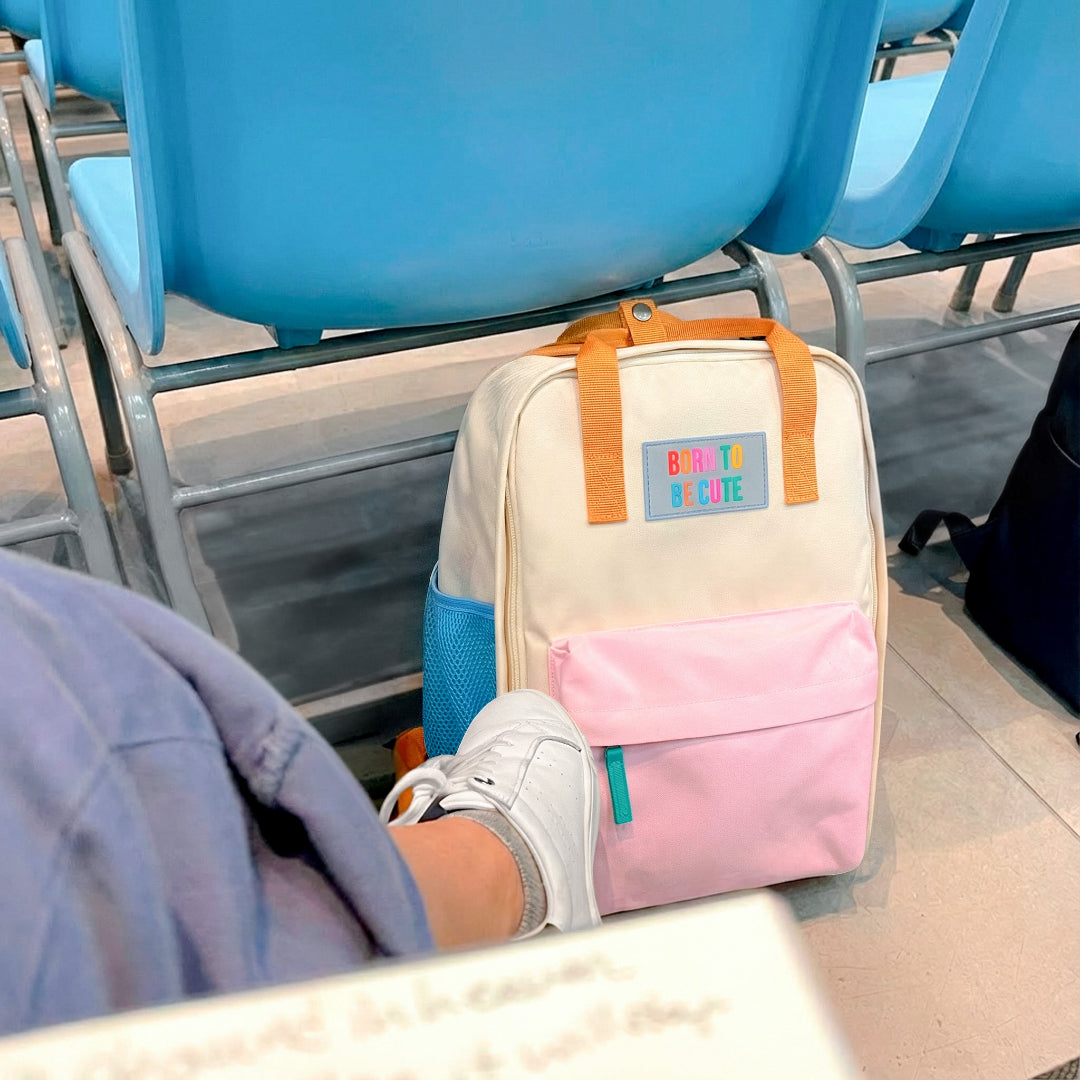 Mochila crema y pastel pequeña con bolsillos y la frase "Born to be cute" bordada en el suelo de una sala de espera