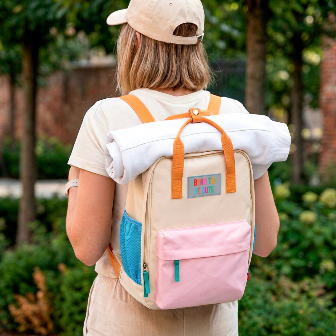 Mujer de espaldas con ropa de deporte y la Mochila crema y pastel pequeña con bolsillos y la frase "Born to be cute" bordada con una sudadera enrollada cogida con las asas