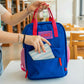 Mujer en la biblioteca metiendo una cartera en el bolsillo delantero de una mochila pequeña azul y roja con el mensaje ciao