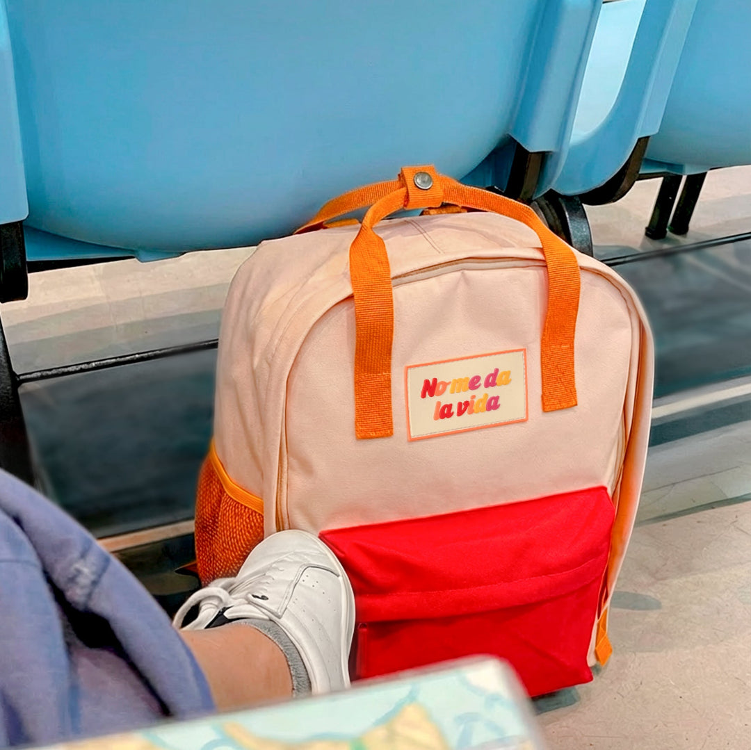 Mochila naranja pequeña con bolsillo y la frase "No me da la vida" bordada en el suelo de un tren