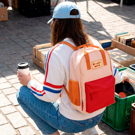 Chica agachada en el rastro con un café en la mano y una mochila naranja pequeña con bolsillo y la frase No me da la vida bordada