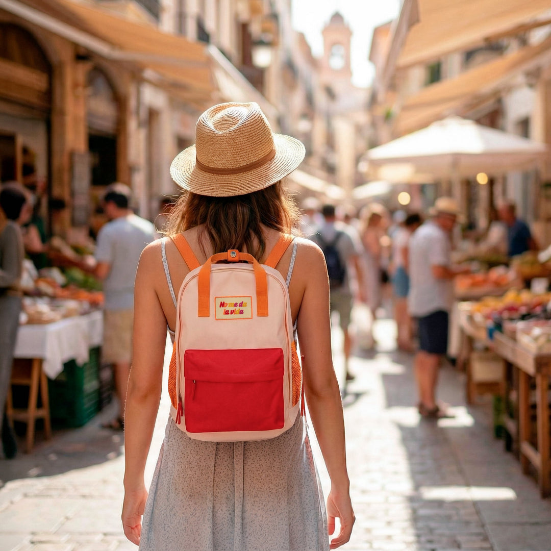 Chica de espalda en un mercado de frutas y verduras con sombrero de paja y mochila naranja pequeña con bolsillo y la frase "No me da la vida" bordada