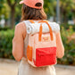 Chica de espalda con gorra y una mochila naranja pequeña con bolsillo y la frase "No me da la vida" bordada y una sudadera enrollada sujeta con las asas