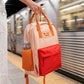 Persona en una estación de tren sujetando la mochila naranja pequeña con bolsillo y la frase "No me da la vida" bordada y una botellita de agua en el bolsillo lateral