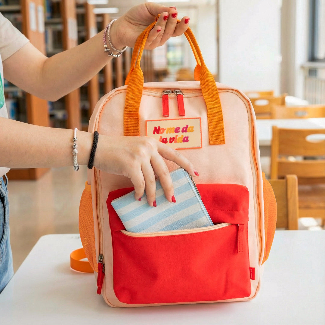 Mujer en una biblioteca metiendo una cartera en el bolsillo delantero de la mochila naranja pequeña con bolsillo y la frase "No me da la vida" bordada