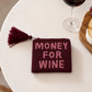 Monedero de cuentas color burdeos con el texto MONEY FOR WINE en rosa y una borla en la cremallera puesto en una mesa junto a una copa de vino tinto y una tabla de quesos