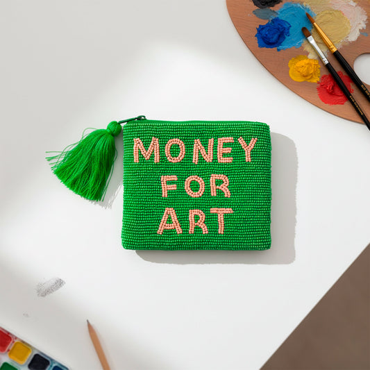 monedero pequeño con cuentas de color verde y el texto "money for art" en cuentas de color naranja. con una borla en color verde en una mesa blanca junto a una paleta de oleos y acuarelas