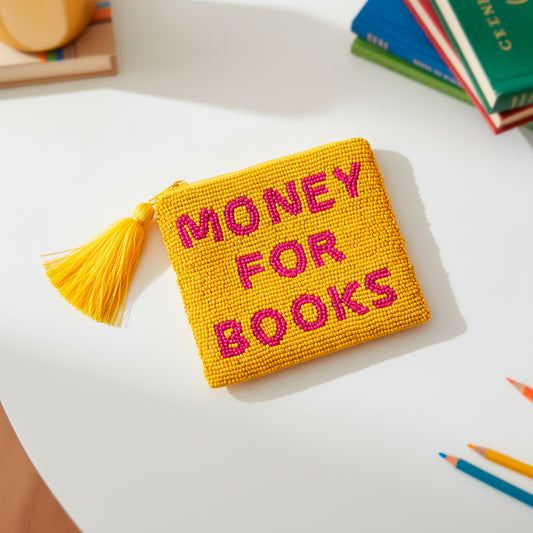 monedero pequeño hecho con cuentas de cristal amarillas y con el texto "money for books" en cuentas de color rosa. Con una borla de color amarillo colgada de la cremallera encima de una mesa blanca junto a lápices y libros