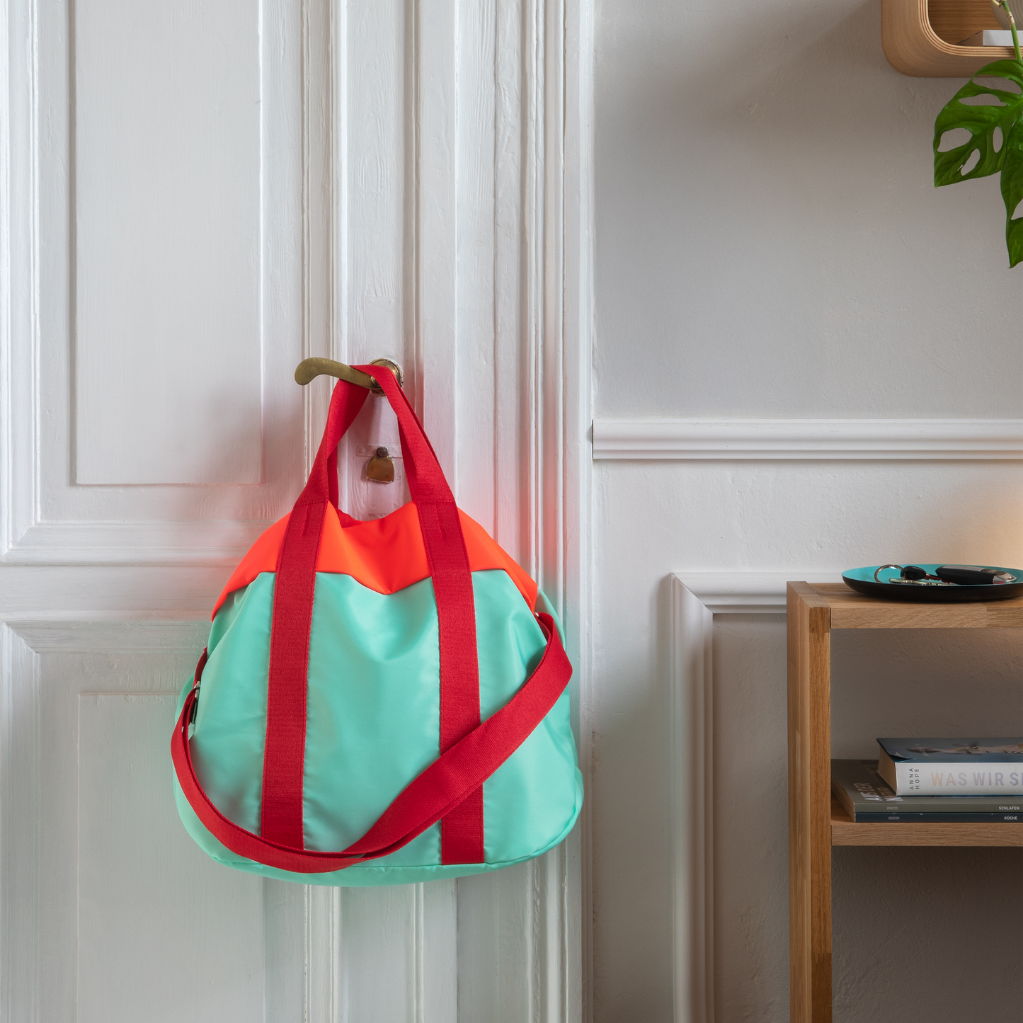 Bolsa de la marca Remember, modelo Nelly en color turquesa con detalles naranjas y correa roja colgada sobre una puerta. 