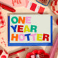 Minitarjeta de cumpleaños One Year Hotter de Laura Ortiz, con letras multicolor sobre fondo claro y sobre azul, entre artículos de papelería.
