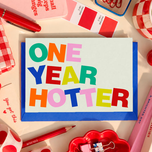 Minitarjeta de cumpleaños One Year Hotter de Laura Ortiz, con letras multicolor sobre fondo claro y sobre azul, entre artículos de papelería.
