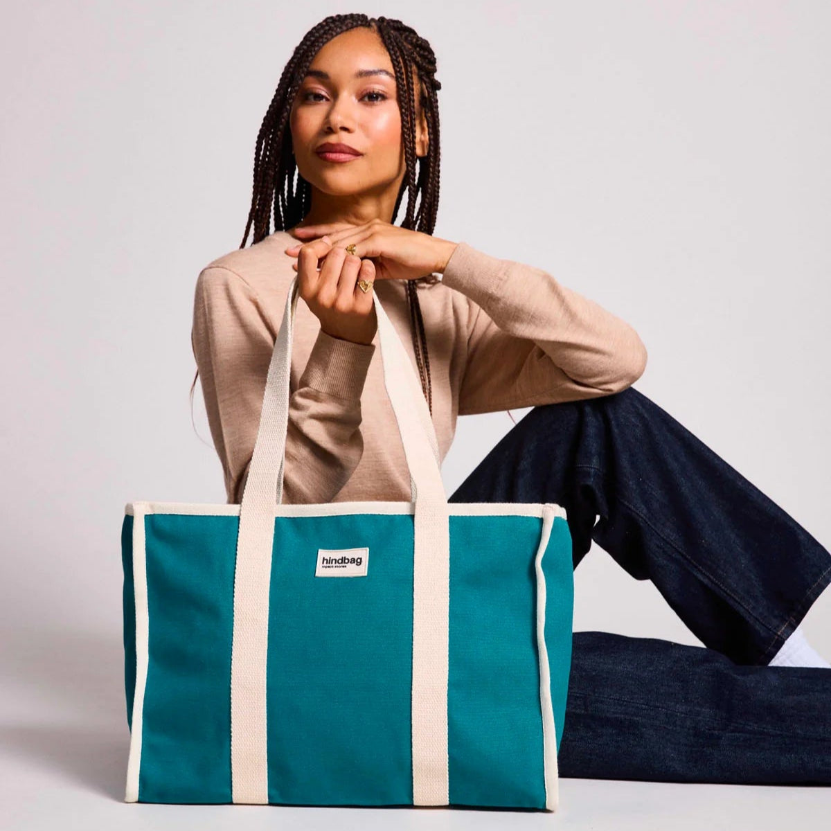 Mujer sentada en el suelo con el bolso grande y cuadrado estilo tote bag de color azul turquesa con asas beige para llevar al hombro de la marca Hindbag