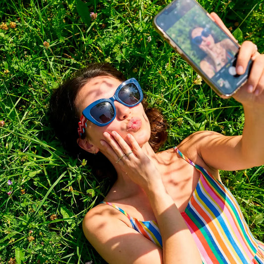chica tumbada en el césped con las gafas puestas de Fisura en color azul con las patillas a franjas rosas y rojas en zig zag del modelo oui cest moi, haciéndose un selfie