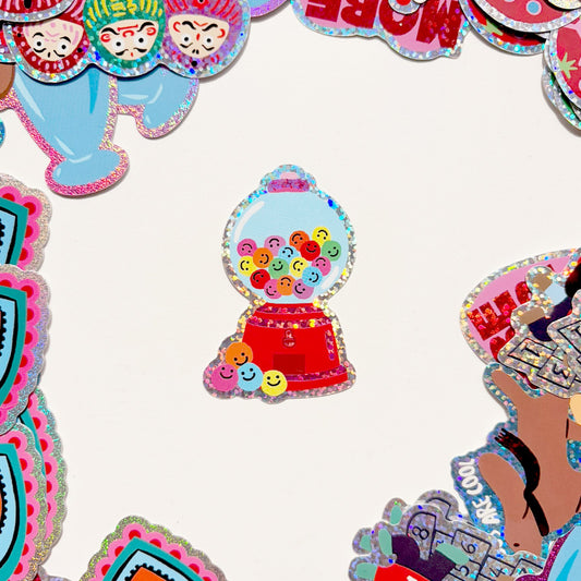 Mano sosteniendo una pegatina con brillo de una máquina expendedora de chicles roja, con bolas de colores con caritas sonrientes, sobre fondo verde claro