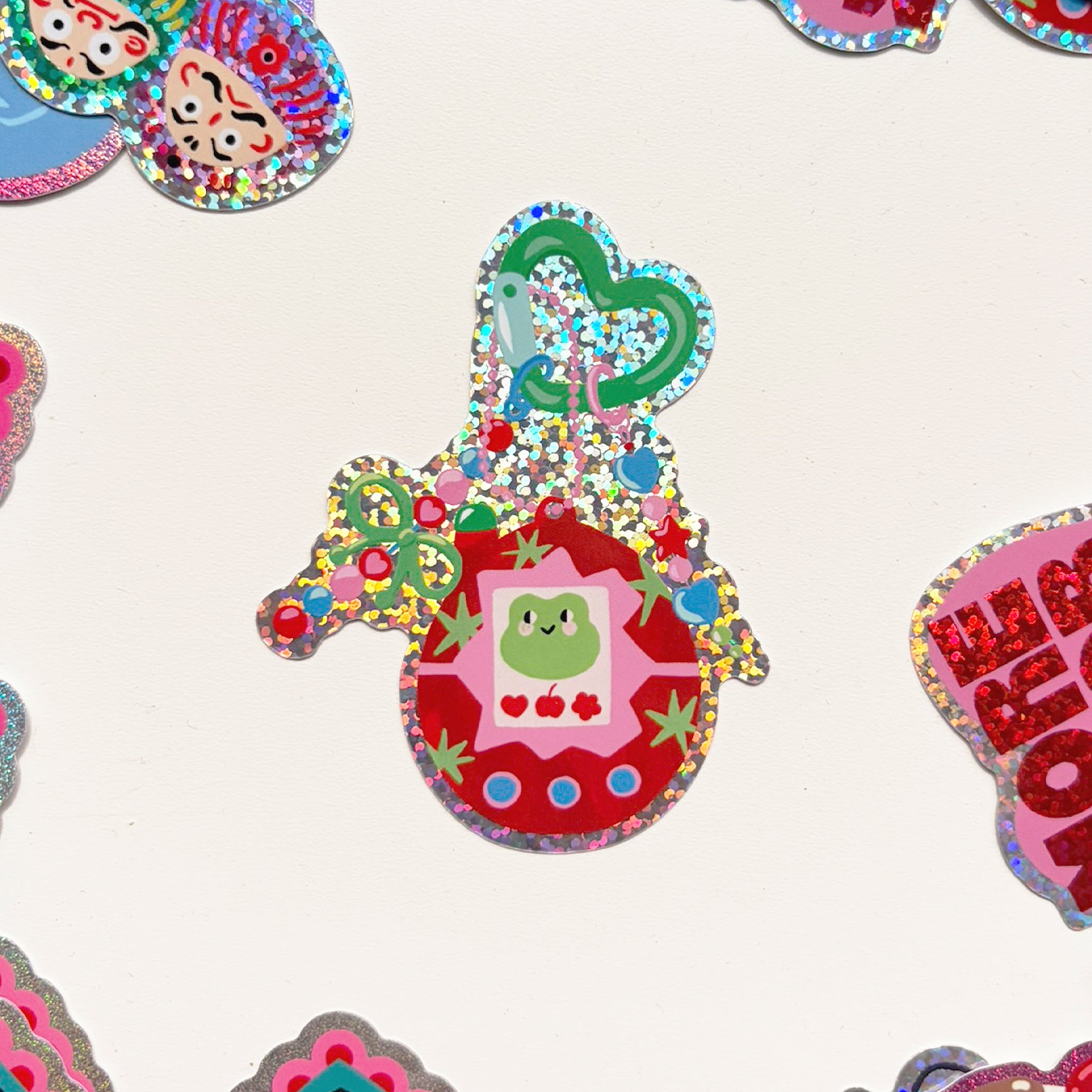 Pegatina con forma de tamagochi con imagen de una rana y bordes de purpurina