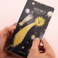 Manos sosteniendo la agenda pocket Astro 2027 de Happimess con portada negra, estrella fugaz dorada y sol con cara, fondo rosa claro