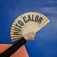 abanico de la marca fisura con purpurina dorada y el texto "PUTO CALOR" en color negro sujetado por una mano y sobre fondo azul y rosa geométricos de la tienda GNOMO